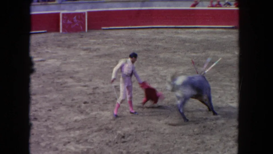 1962 Los Angeles home-movie frame showing a matador presenting a red capote to a grey bull in a sandy arena.