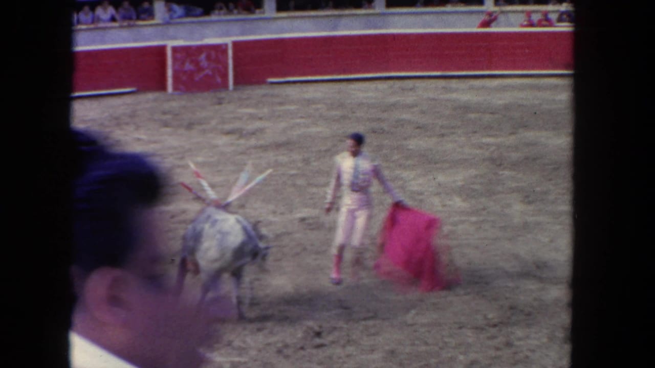 1962 Los Angeles arena footage with a grey bull, matador, red capote, crowd seating, and a blurred spectator crossing the foreground.