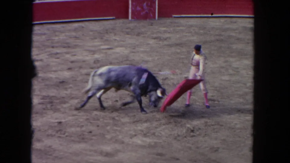 The matador draws the red cloth away while the grey bull lowers its head on the dusty arena floor in this Los Angeles 1962 frame.