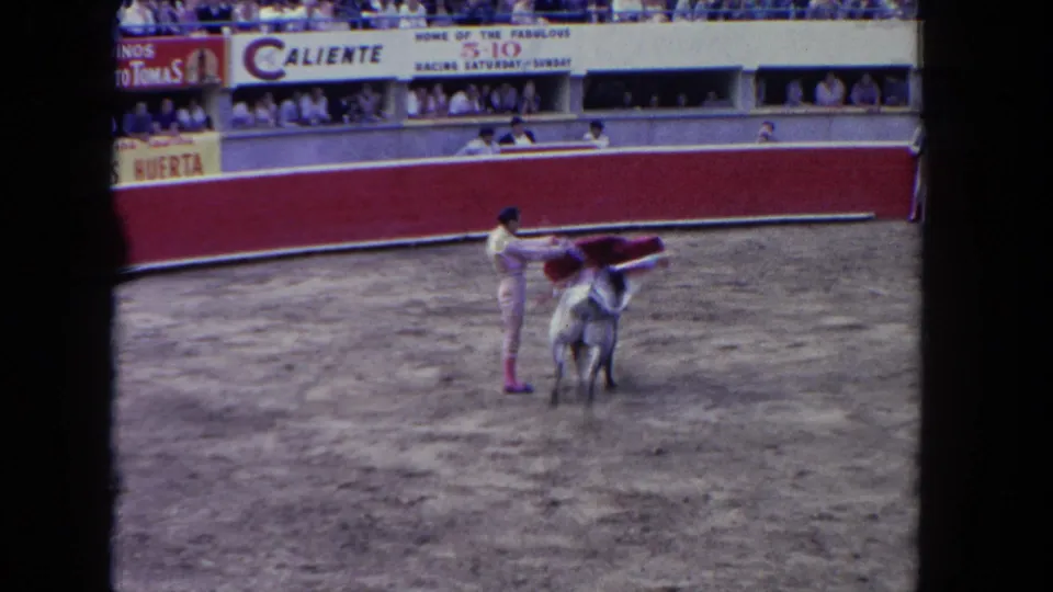 1962 Los Angeles 8mm frame with a matador maneuvering a red cape near a bull in front of arena seating and signage.