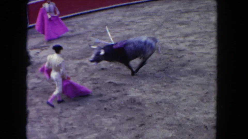 1962 Los Angeles 8mm frame showing a bull facing matador-costumed participants with bright pink capes in the dirt arena.