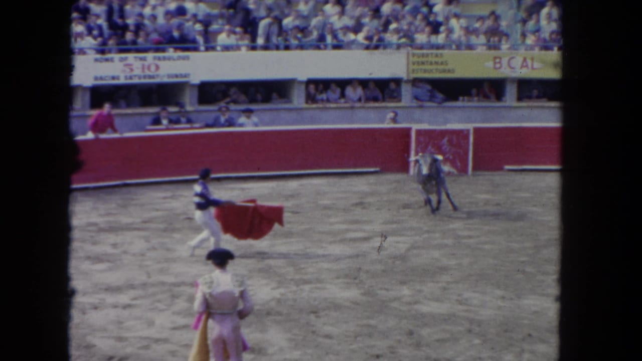 1962 Los Angeles home-movie frame showing a bullfighter holding a red cape as a bull moves across a sandy arena before seated spectators.