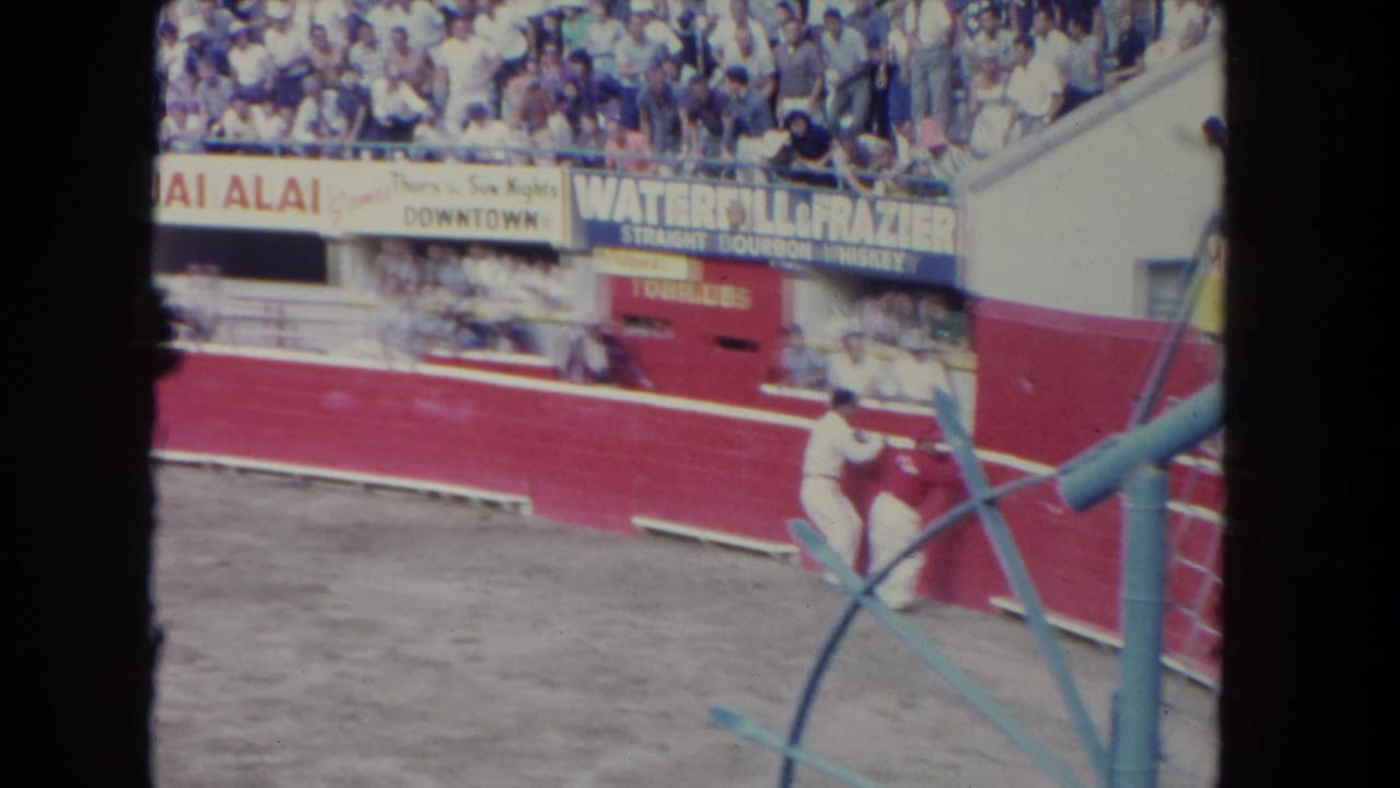 Matador near red arena wall as a bull charges, with crowded stands and painted signs behind.