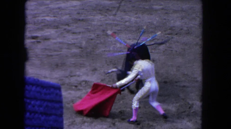 1962 Los Angeles home-movie frame showing a matador in a light suit holding a red cape near a black bull in a bullring.