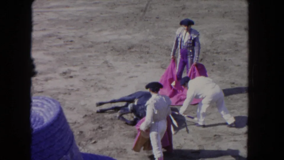 1962 Los Angeles 8mm frame showing three matador-like figures with pink capes near a bull lying on the sandy arena floor.