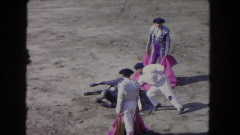 1962 Los Angeles 8mm frame showing a man bending beside the downed bull as two others hold capes nearby in the arena.