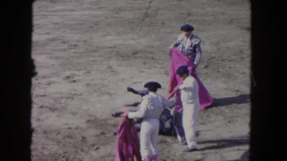 1962 Los Angeles 8mm frame showing capes gathered around a bull on the ground as matador-like figures stand in the sandy arena.