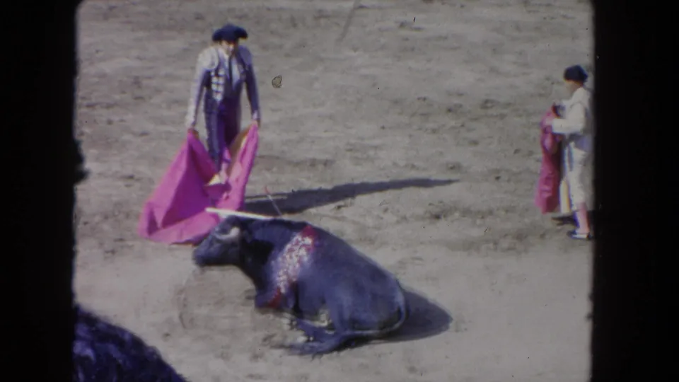 1962 Los Angeles arena footage shows a bull on the ground, a caped performer nearby, and another participant holding cloth at right.