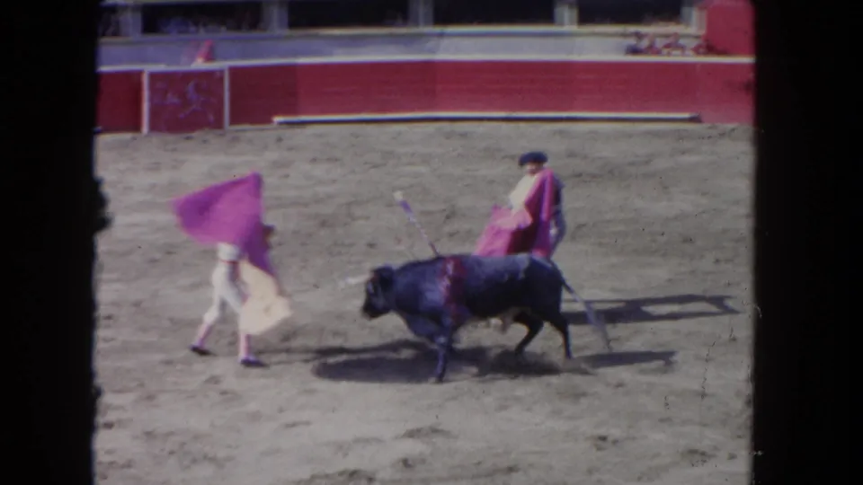 1962 Los Angeles 8mm frame showing two matadors in a dusty ring as a black bull moves between their pink capes.
