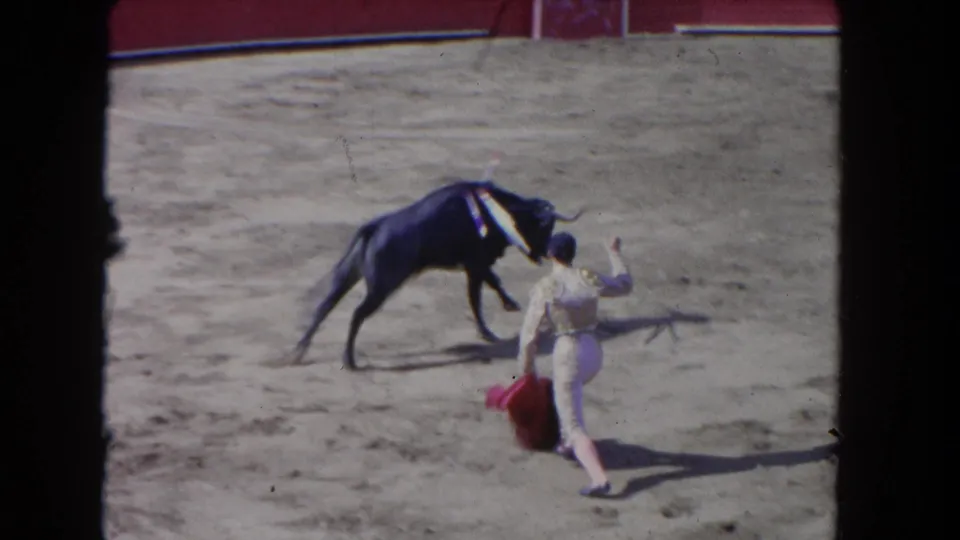 The bull lowers its head and moves toward the bullfighter, who steps away with the red cape in this 8mm arena footage.
