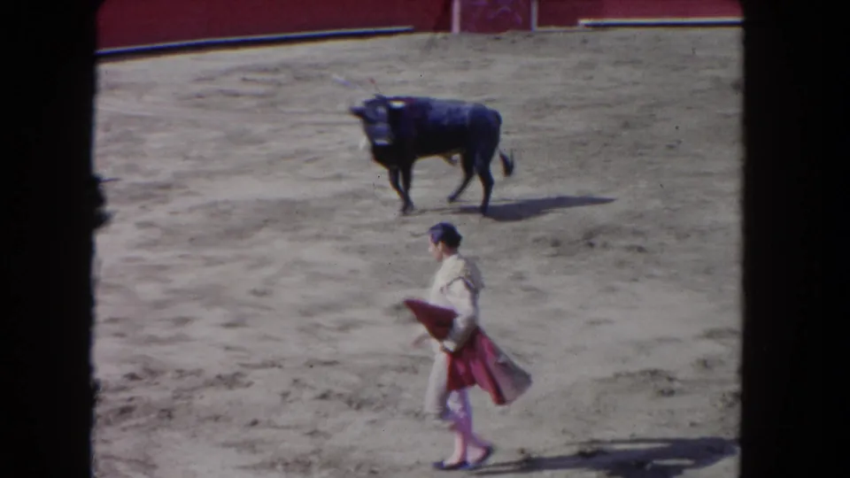 The bullfighter carries the red cape across the sandy arena while the dark bull stands at a distance in the 1962 Los Angeles clip.