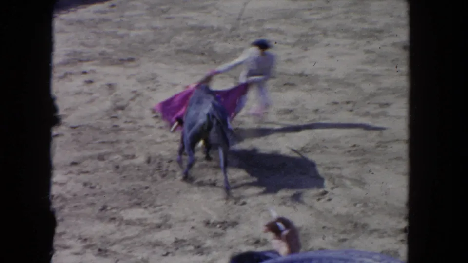 1962 Los Angeles 8mm frame showing a white-clad matador-style performer using a red cape near a dark bull in a sunlit arena.