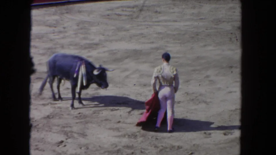 1962 Los Angeles 8mm frame showing the performer from behind, holding a red cape while facing a ribboned bull in the arena.