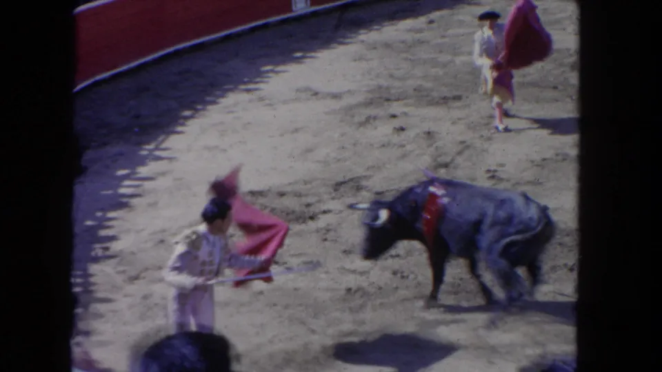 1962 Los Angeles 8mm frame showing a matador with a cape facing a black bull in a sunlit sandy arena.