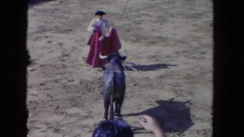 1962 Los Angeles 8mm frame showing a bull moving toward a caped matador, filmed from the stands above the sandy arena.
