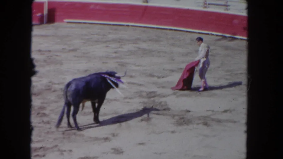 1962 Los Angeles 8mm frame with a black bull facing a matador-style performer holding a red cape in a sandy bull ring.