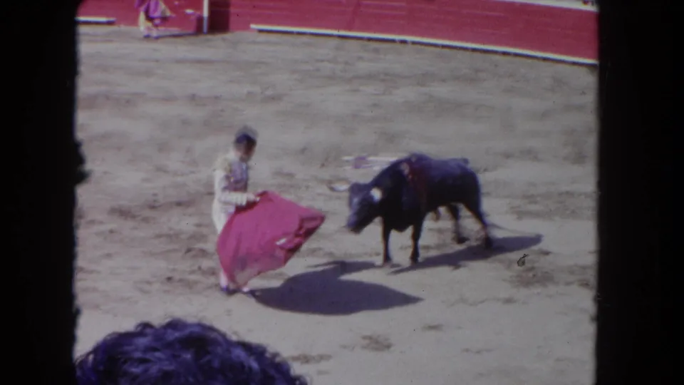 1962 Los Angeles 8mm frame showing a matador-style performer turning a red cape as a dark bull watches in the sandy ring.