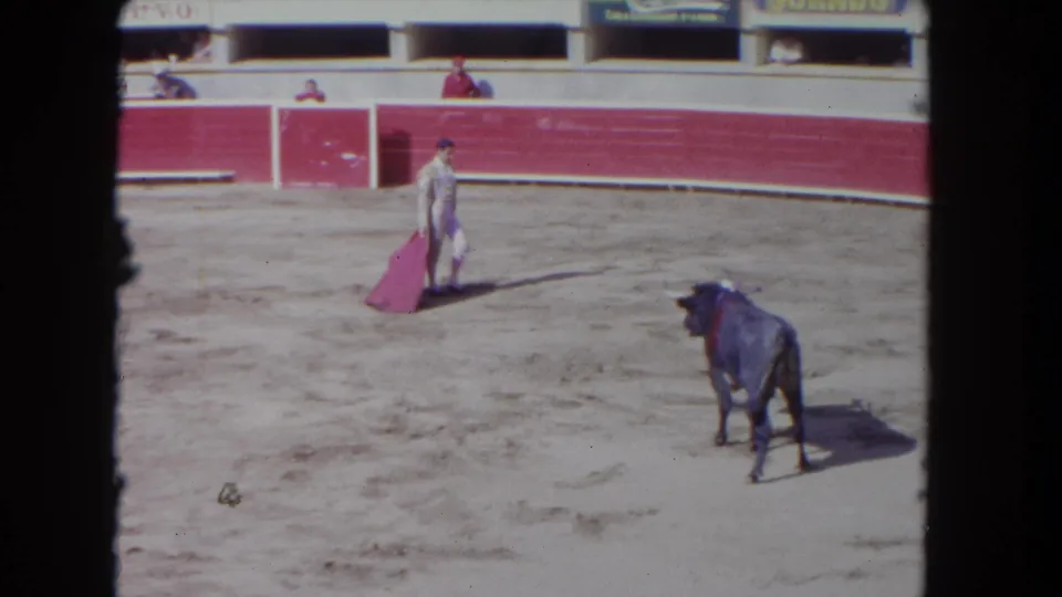 1962 8mm frame shows a matador with a pink cape facing a dark bull inside a sandy bullring, with red barriers and signage behind.