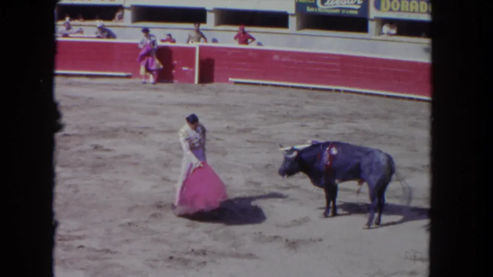 In the 8mm frame, the matador stands close to a dark bull while holding a pink cape low on the sandy bullring floor.