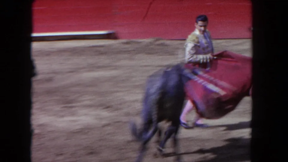1962 Los Angeles home-movie frame showing a matador with a red cape confronting a charging black bull in a sandy arena.