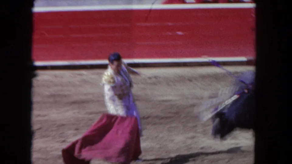 1962 Los Angeles 8mm frame showing a matador with a red cape facing an approaching bull in a dirt-ring arena.