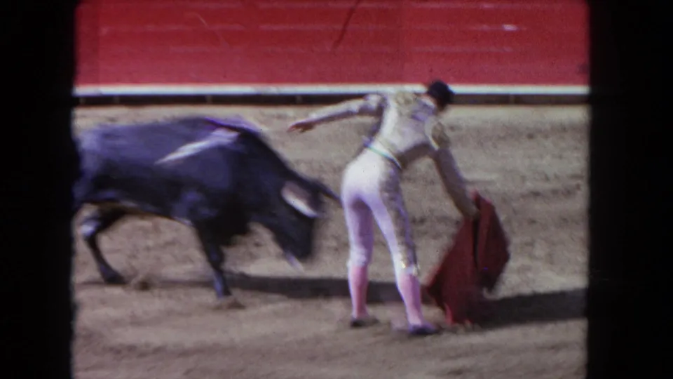 The bull rushes past the matador and red cape in the sandy arena, captured in blurred 8mm motion from the stands.