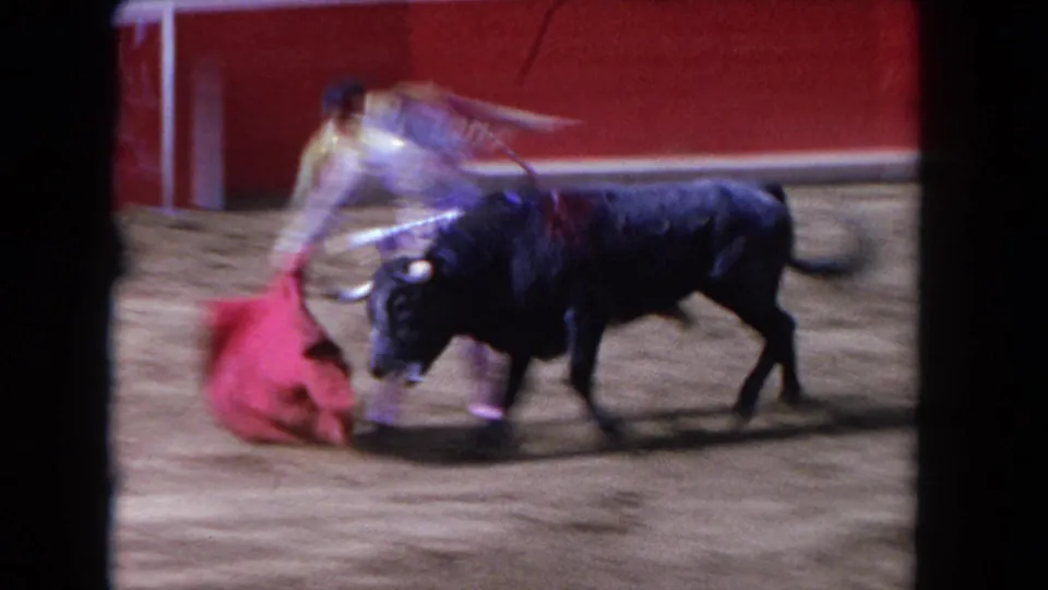 1962 Los Angeles 8mm frame showing a matador moving a red cape as a black bull charges nearby in a sandy ring.