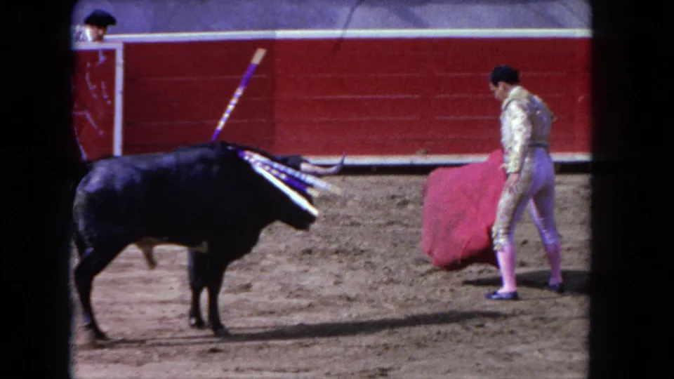 1962 Los Angeles 8mm frame with a black bull facing a matador holding a red cape near red wooden ring boards.
