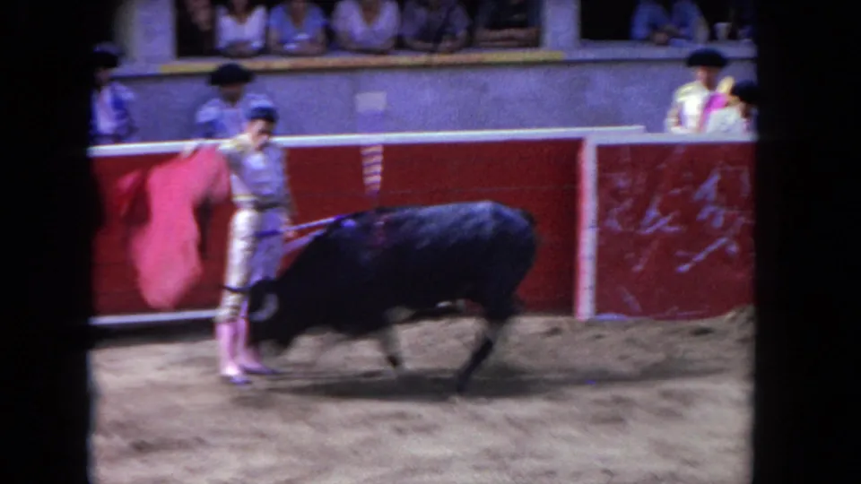 In the 1962 Los Angeles footage, a matador in a light embroidered suit stands near a black bull inside a red-walled bullring.