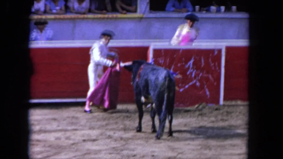 1962 Los Angeles home-movie frame of a matador in a sandy arena facing a black bull with a pink cape as spectators watch behind red boards.