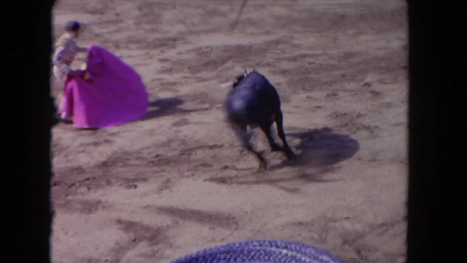 1960s 8mm-style frame of a matador with a bright pink cape facing a black bull in a sunlit bullring.