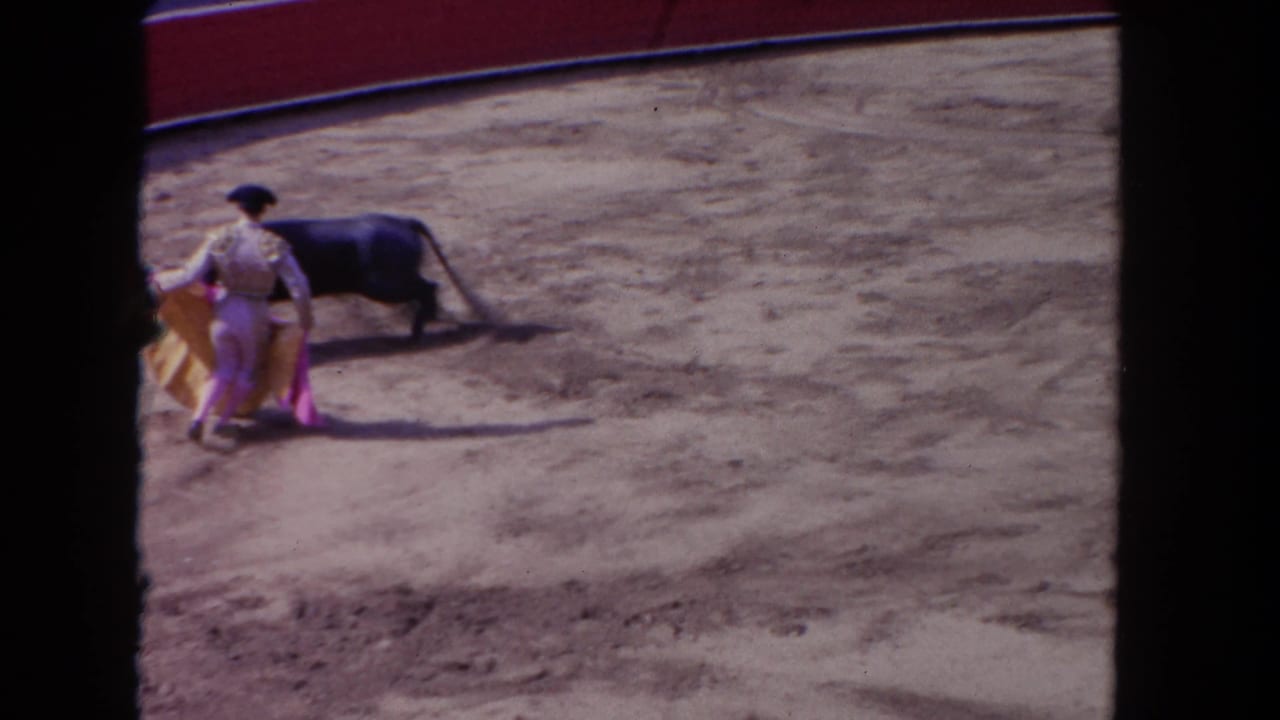 Spectator-shot 8mm frame showing a matador beside a black bull, with the cape lowered on the dirt surface.