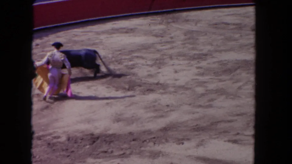 Spectator-shot 8mm frame showing a matador beside a black bull, with the cape lowered on the dirt surface.