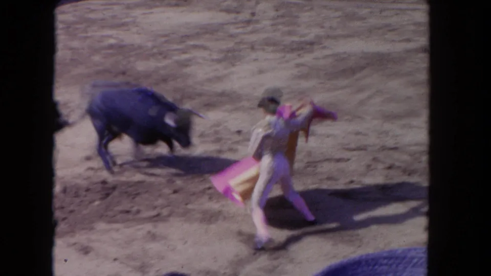 Home-movie frame of a black bull lunging toward a matador who turns with a bright cape on the dirt arena floor.