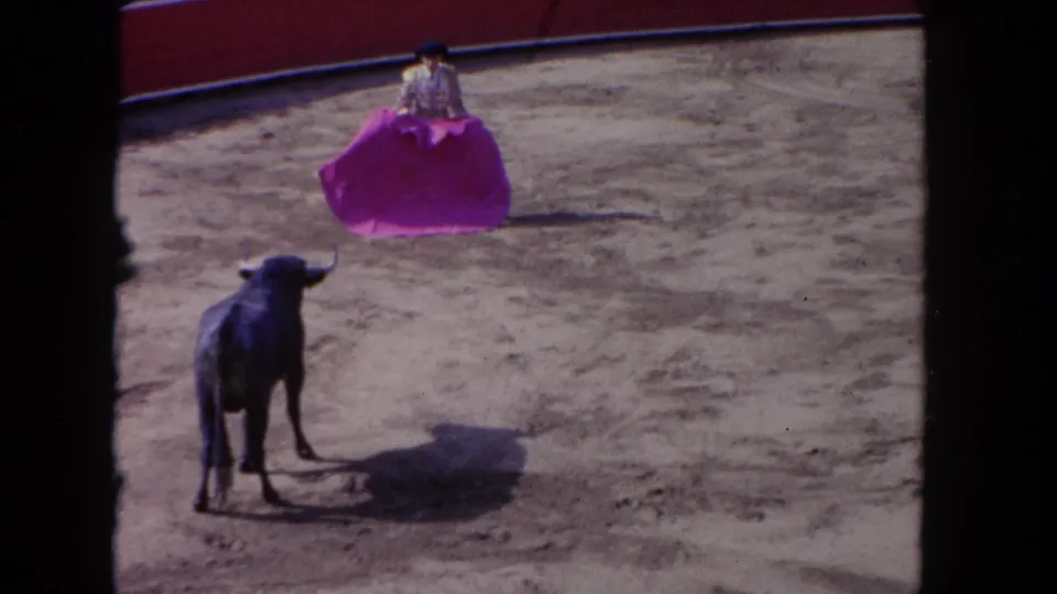 1962 Los Angeles 8mm frame showing a dark bull approaching a torero with a vivid purple cape inside a sandy bullring.