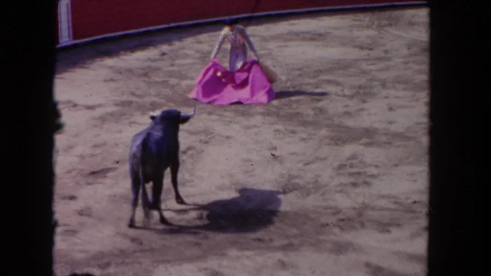 1962 Los Angeles home-movie frame with a black bull in the foreground and a matador holding a pink cape in the arena.