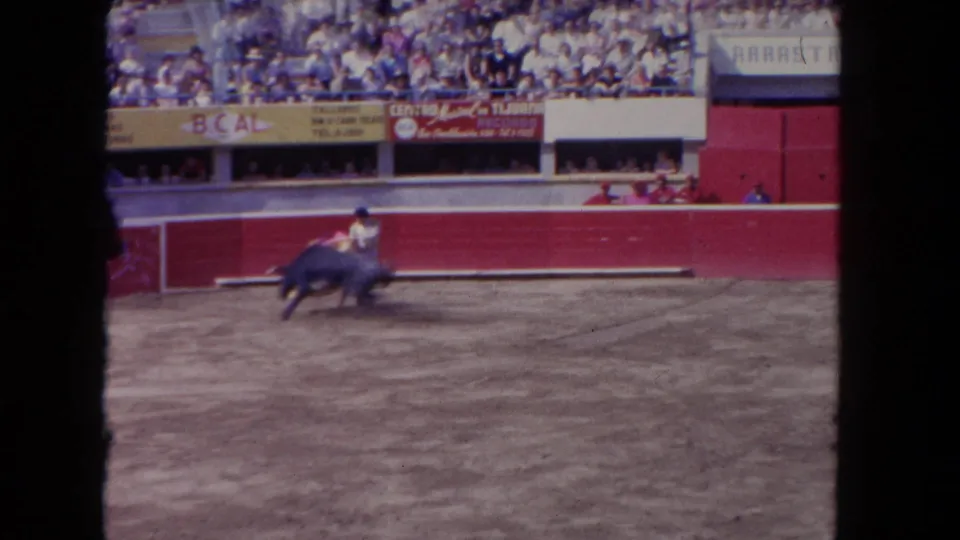 1962 Los Angeles 8mm frame showing a matador near a charging dark bull, with red arena walls and spectators behind.