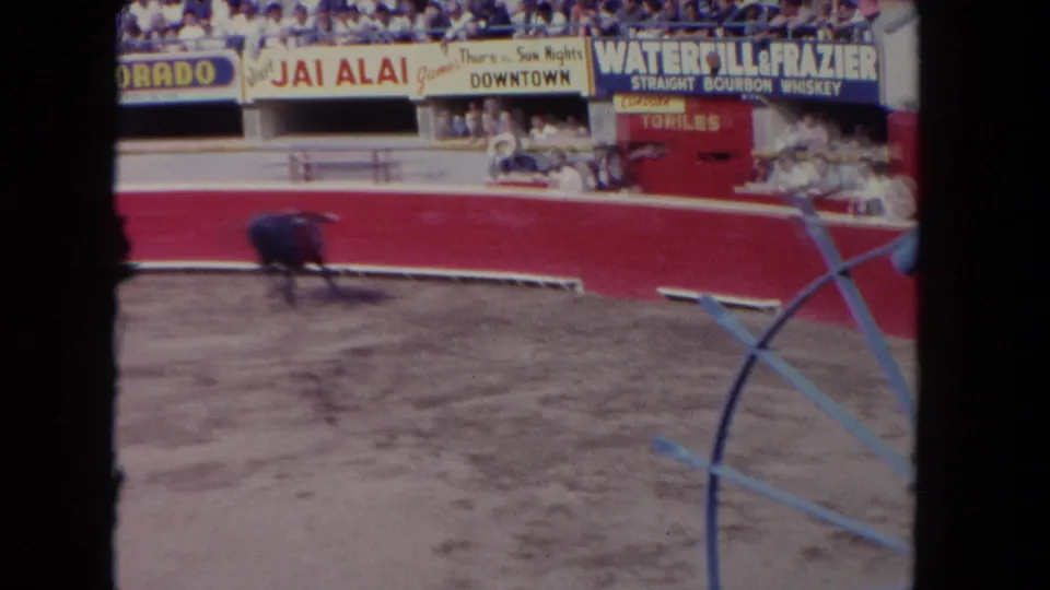 1962 Los Angeles home-movie frame showing a dark bull moving across a sandy arena with red barriers, spectators, and arena ads behind it.