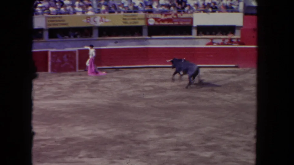 1962 Los Angeles home-movie frame showing a matador holding a pink cape as a dark bull charges across the arena before spectators.