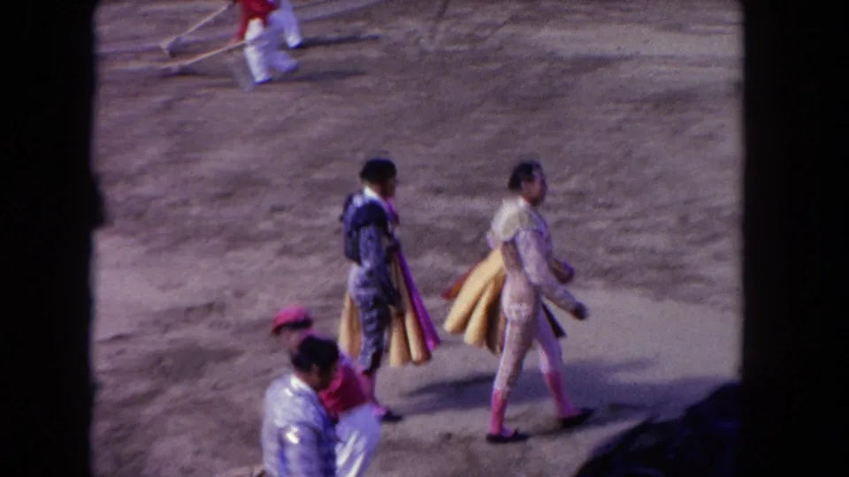 1962 Los Angeles 8mm frame showing matadors in ornate costumes carrying capes across a dusty bullfighting arena.