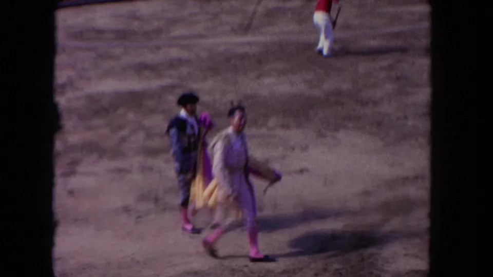 1962 Los Angeles 8mm frame showing matadors in suits of lights crossing the bullring as workers sweep near the red barrier.