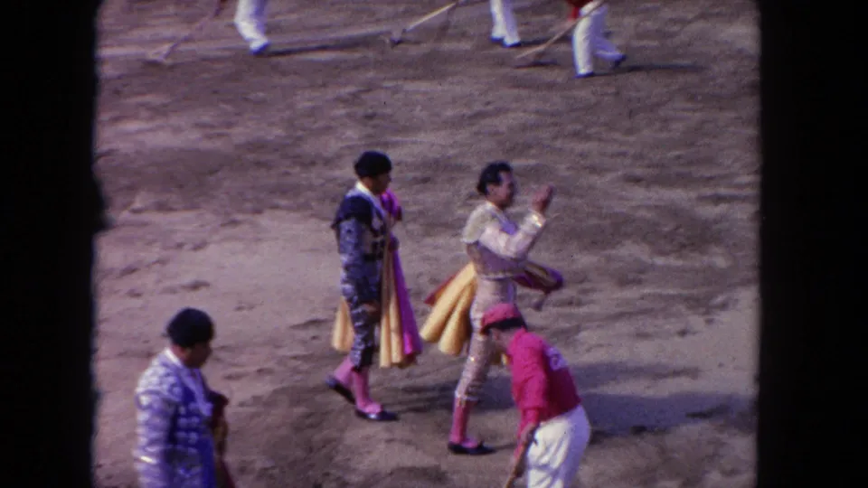 1962 Los Angeles 8mm frame with matadors walking across the arena, a worker in red, sweepers in back, and spectators above red barriers.