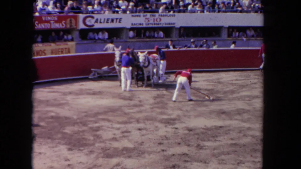 At a 1962 Los Angeles racetrack, a red-shirted worker rakes the dirt as other workers tend horses near the rail.