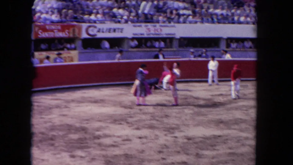 1962 Los Angeles 8mm frame showing workers and matadors in a dirt bullring with a crowd and advertising signs behind a red barrier.