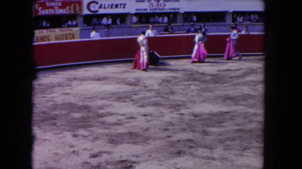 1962 Los Angeles arena scene with matador figures in white and pink near a red barrier as spectators watch from the stands.