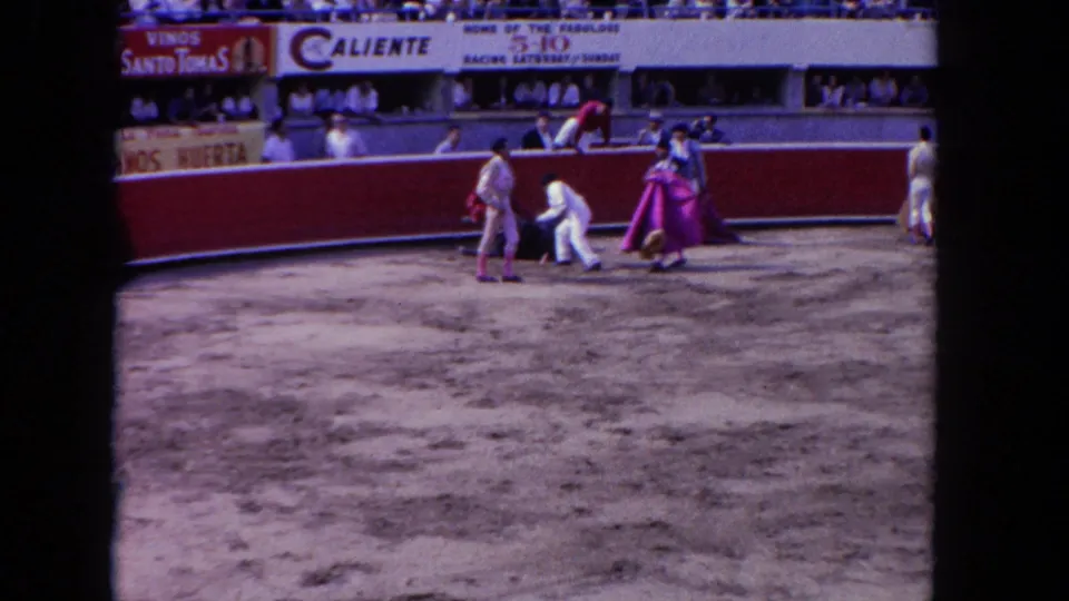 Wide 1962 Los Angeles arena view with matador-style performers, capes, spectators, signage, and one figure bending near the ground.