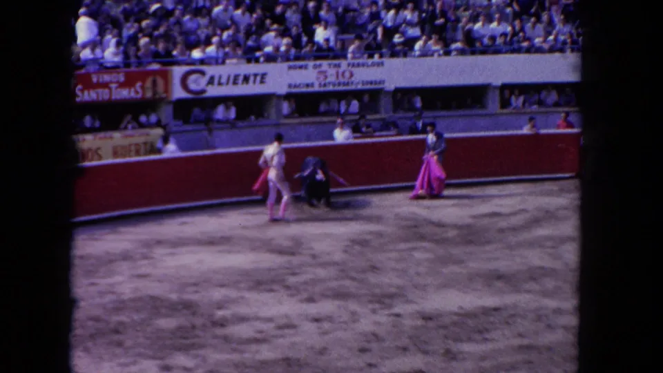 A 1962 home-movie view of a matador drawing a black bull toward a cape near the barrier, with a packed crowd behind the ring.
