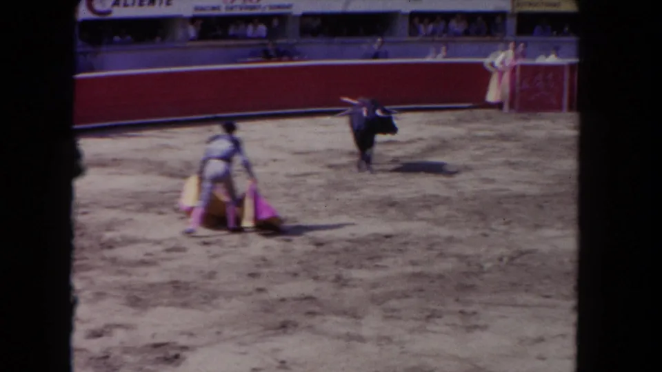 1962 Los Angeles 8mm frame showing a matador with cape facing a black bull in a sunlit arena as spectators watch from the stands.