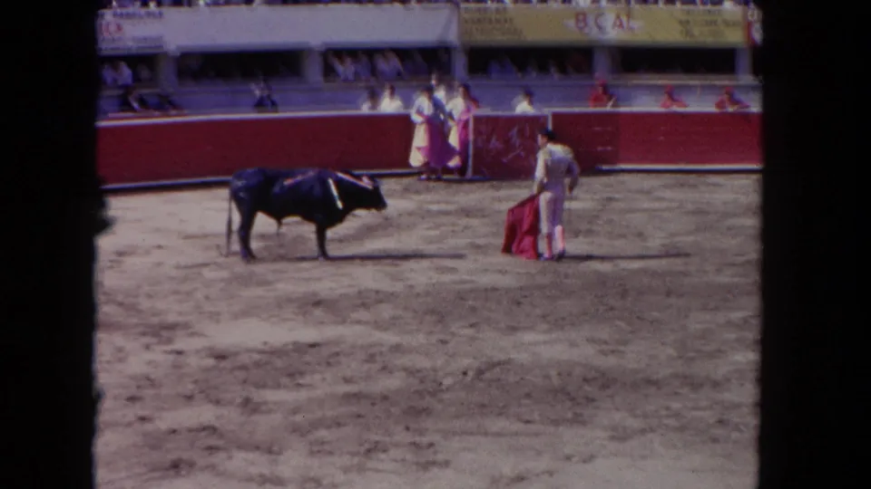 1962 Los Angeles 8mm frame showing a matador and black bull separated across a dusty arena with spectators behind the red wall.
