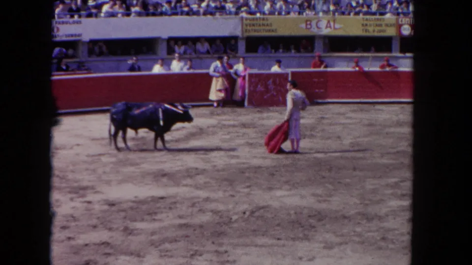 1962 Los Angeles 8mm frame showing a wide bullfighting arena view with bull, matador, red cape, spectators, and signage above the barrier.